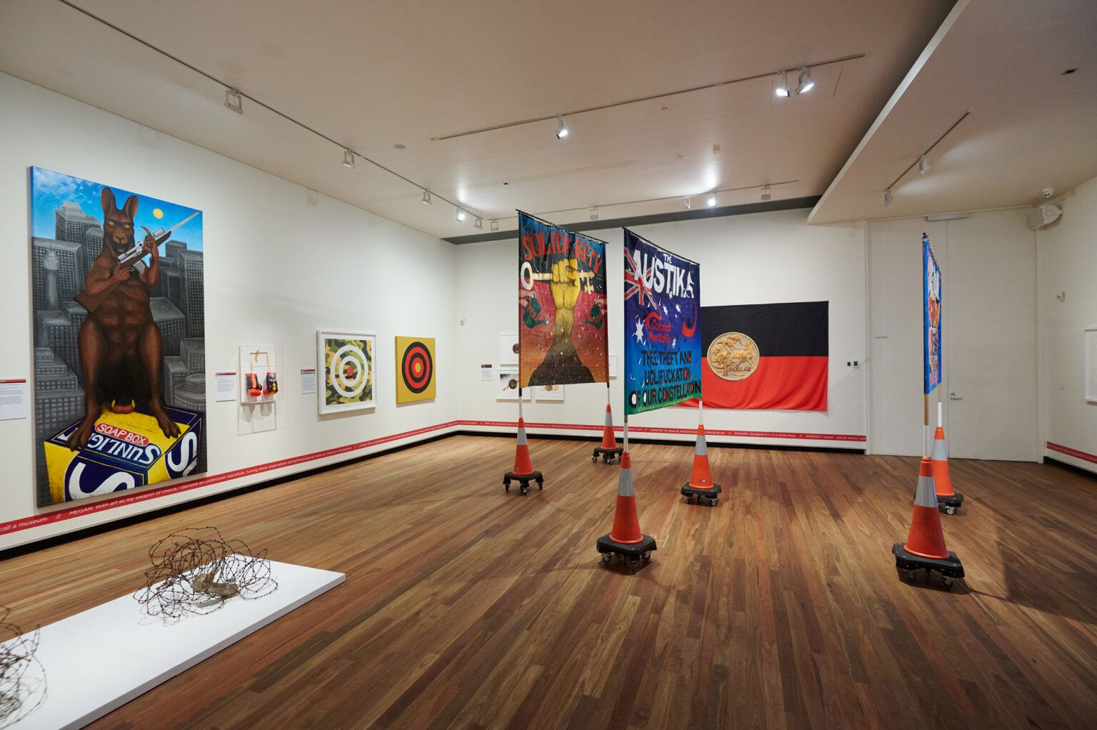 A view of an exhibition featuring a collection of artworks including banners, flags and paintings in a white room with wooden floorboards. The artworks feature imagery of Australian identity, including the Aboriginal flag.