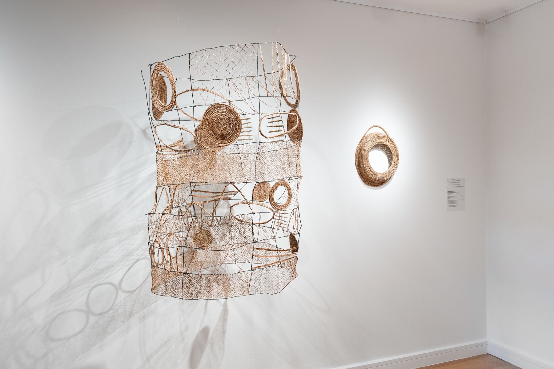Two sculptural artworks made of brown dried reed grass sitting on a white gallery wall.