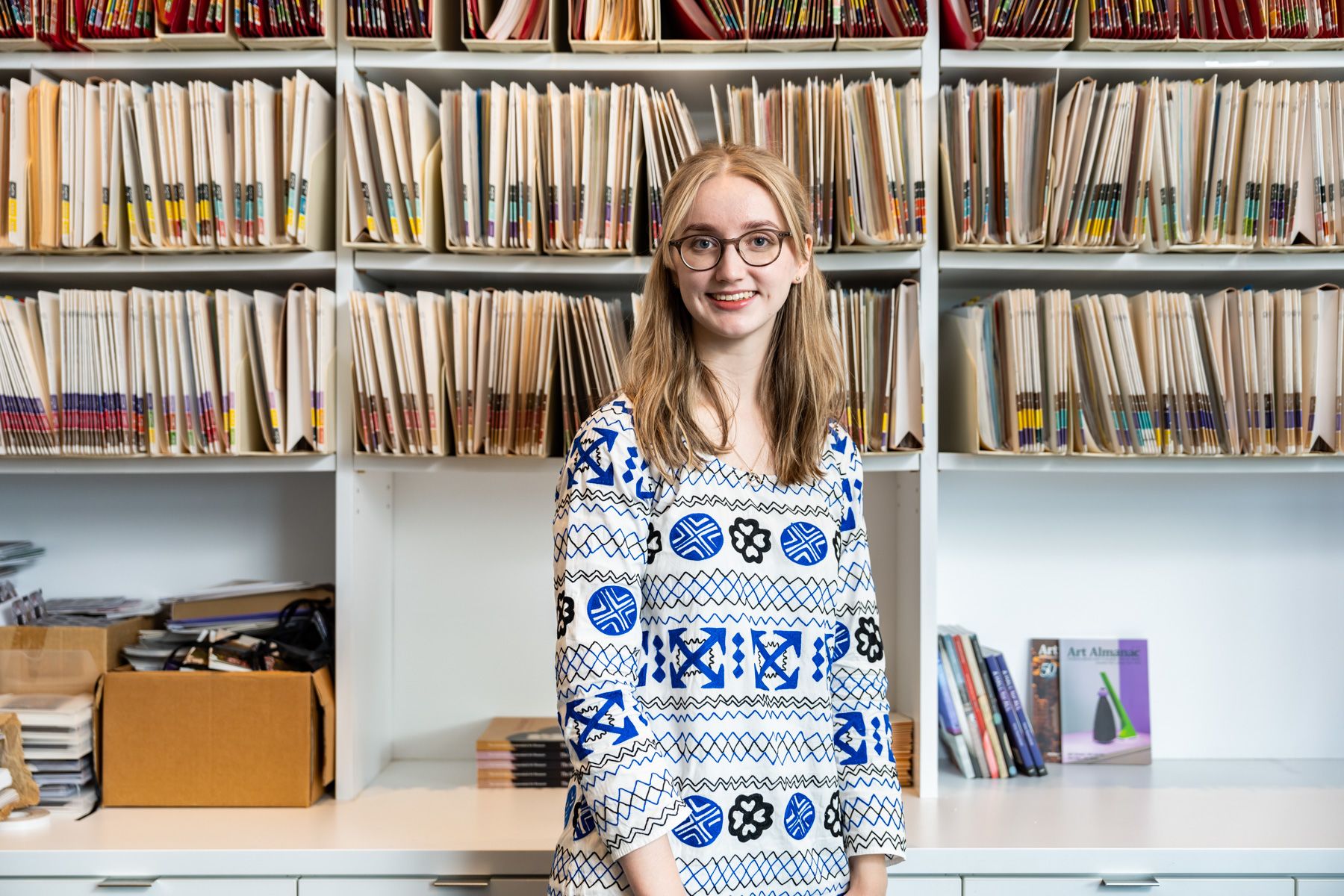 A young person is smiling and standing in front of a shelf filled with manilla folders.