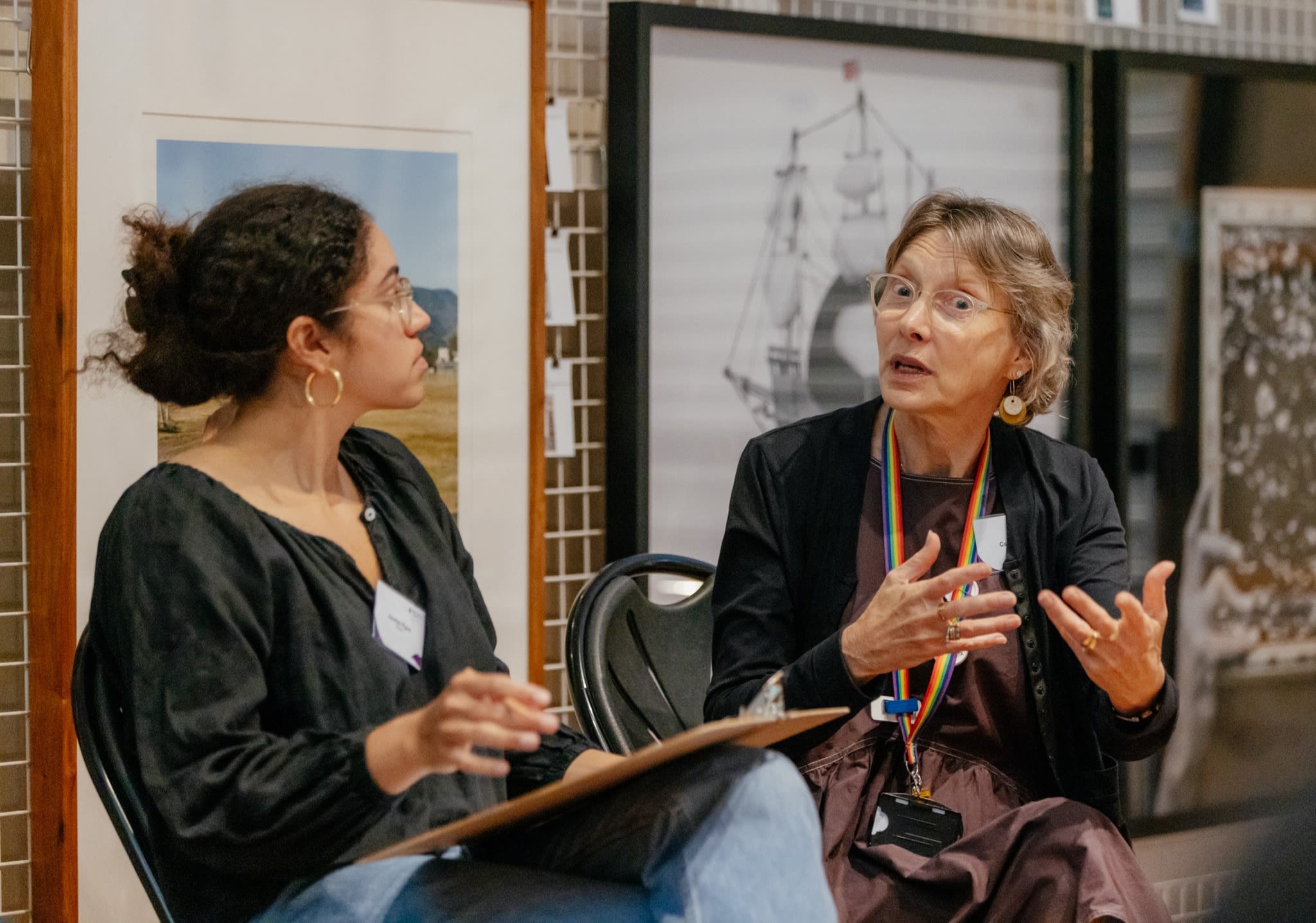 Two people seated in front of artworks. One person is talking and gesturing with their hand whilst the other person is listening.