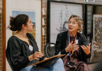 Two people seated in front of artworks. One person is talking and gesturing with their hand whilst the other person is listening. 