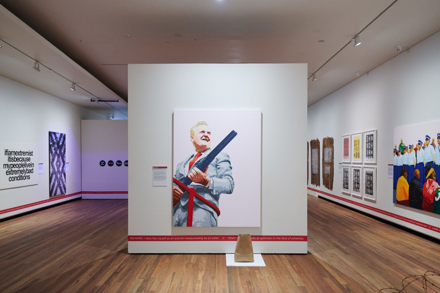 A view of an exhibition in a white walled room. The focus artwork is a painting of a man in a grey suit hold the long barrell of shot gun.