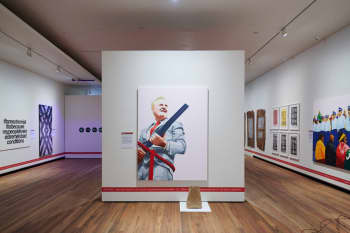 A view of an exhibition in a white walled room. The focus artwork is a painting of a man in a grey suit hold the long barrell of shot gun.