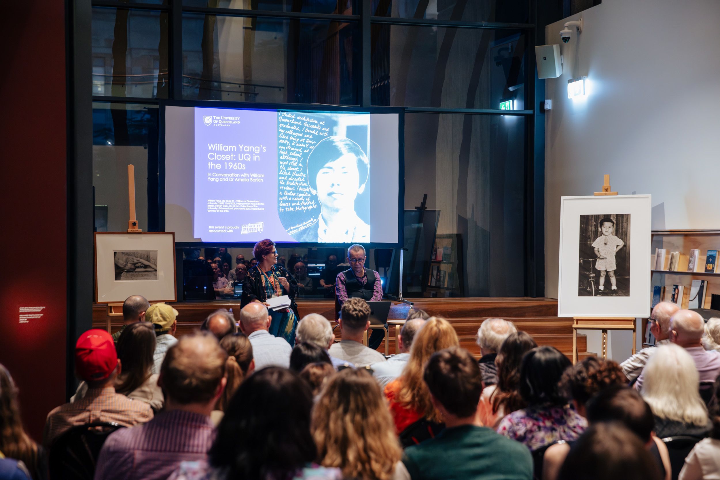 Two speakers are sitting on stools in front of a large seated audience. Behind the speakers there are two photographs on easels and a projection of an image. 