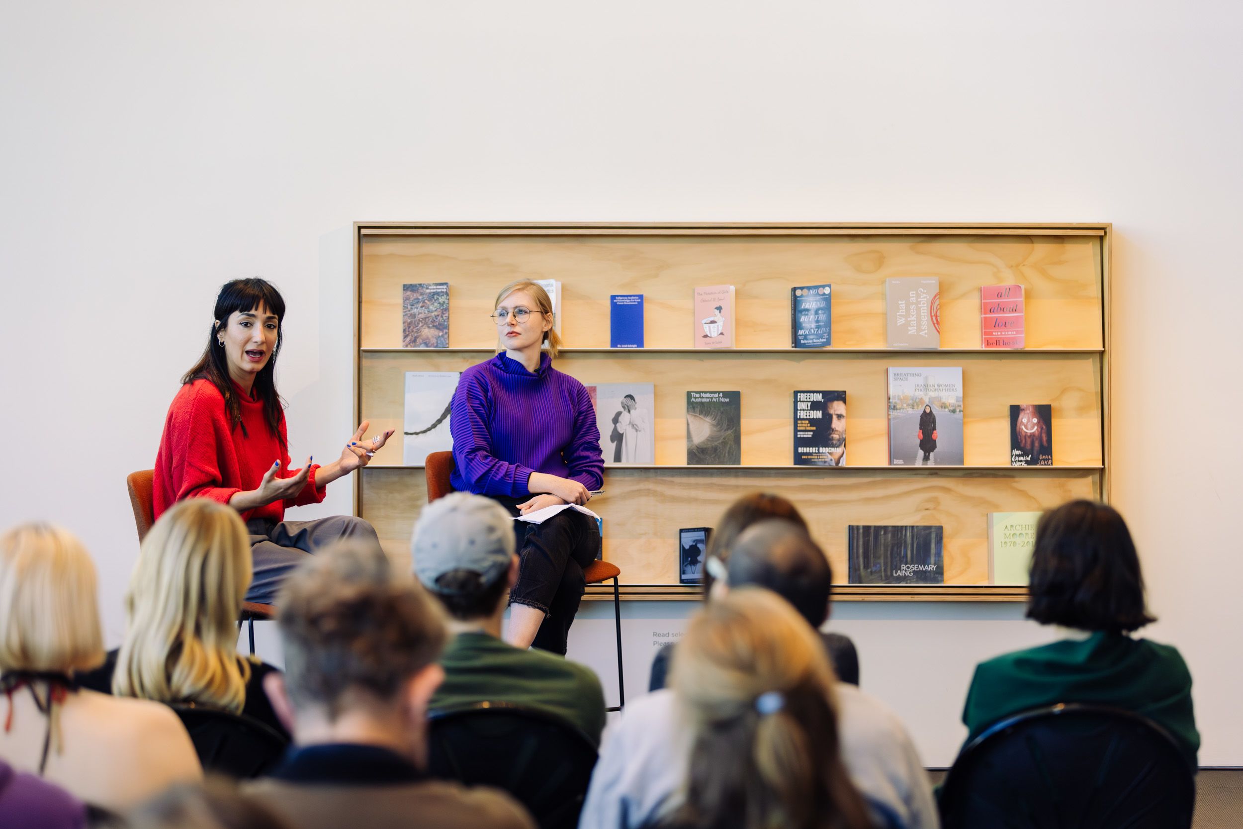 Two people sitting on stool speaking to an audience of seated people. There is a shelf with books displayed on it behind the speakers.