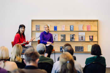 Two people sitting on stool speaking to an audience of seated people. There is a shelf with books displayed on it behind the speakers. 