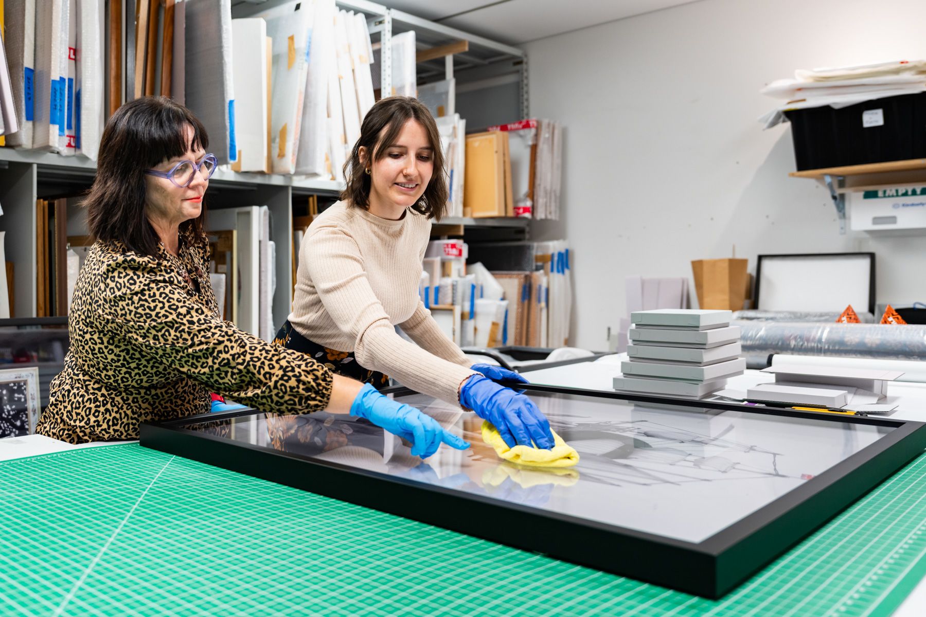 Two people cleaning the glass on a framed artwork in a workshop.