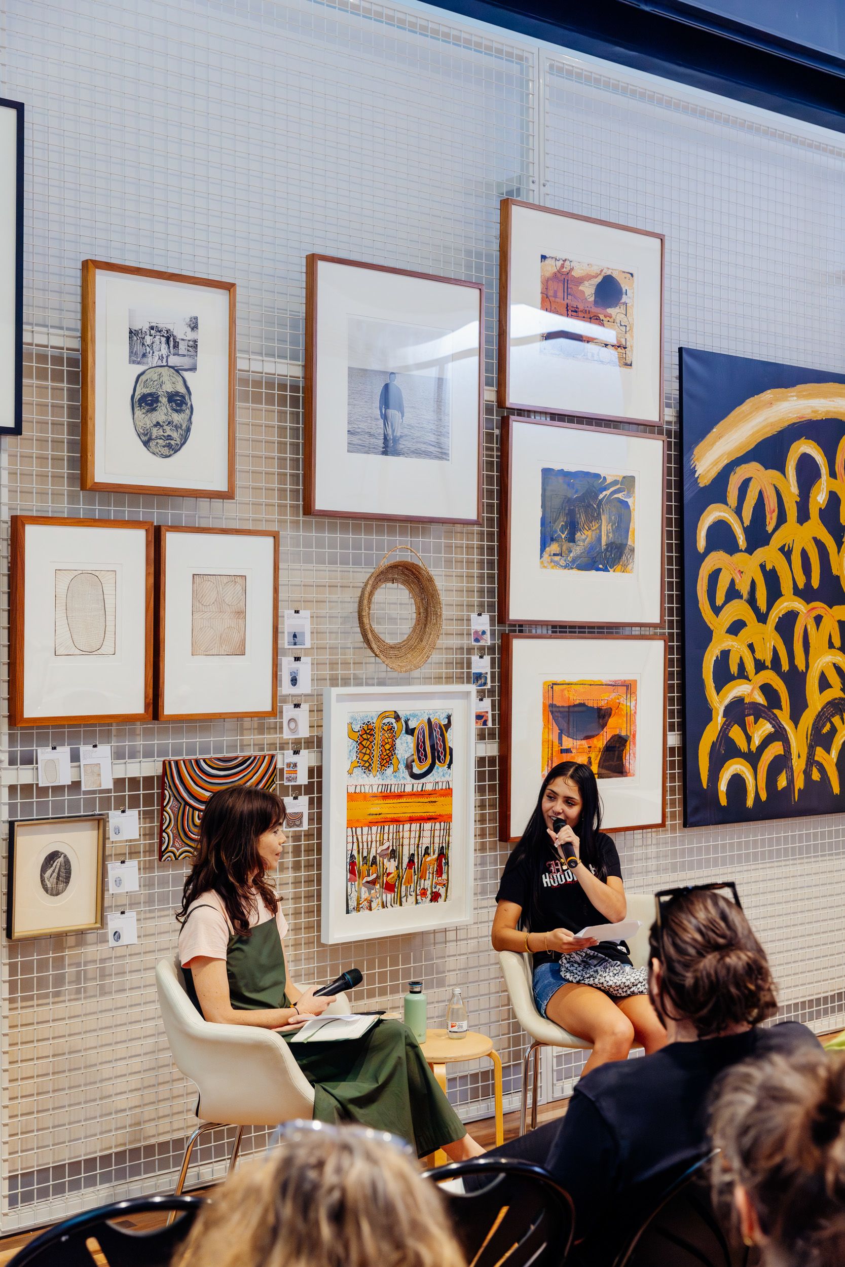 Two people seated in front of artworks. One person is talking in to a microphone and the other person is listening. 