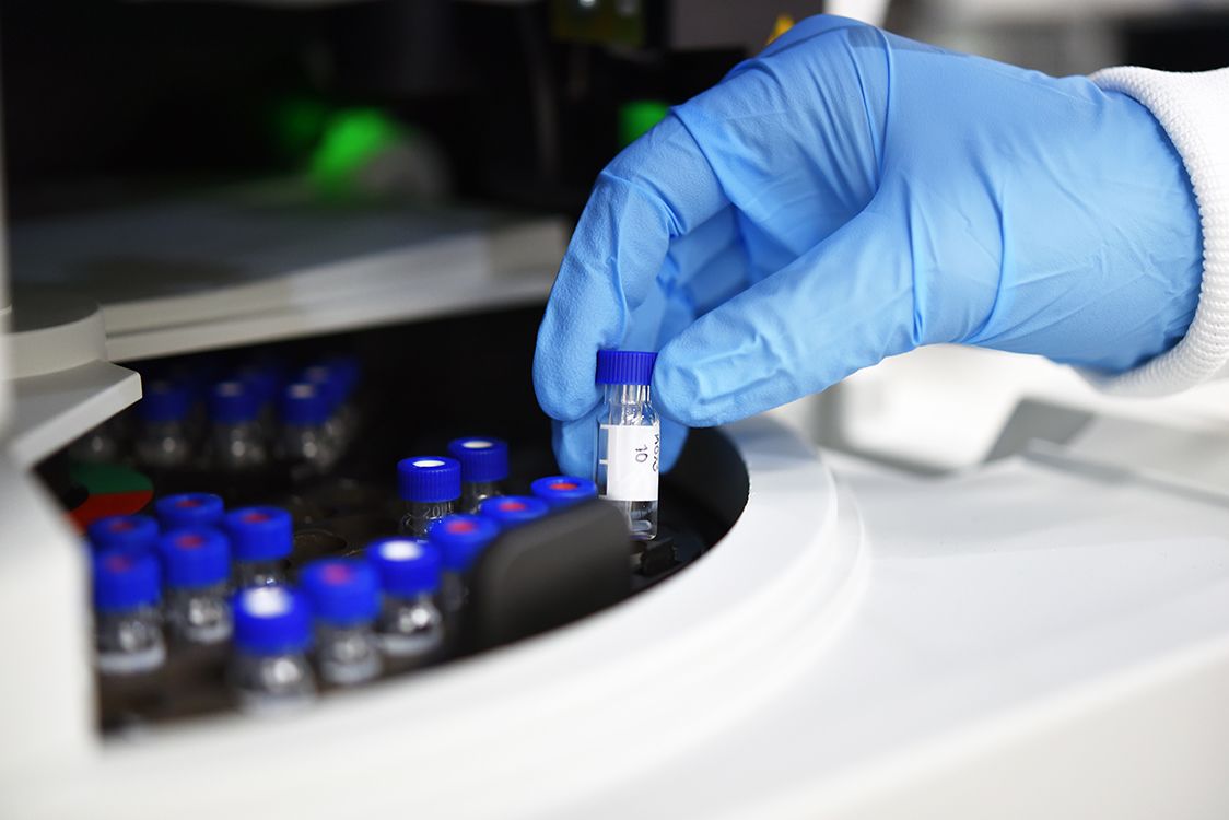 Gloved hand of scientist holding test tube of liquid partially out of a machine with many similar lidded test tubes