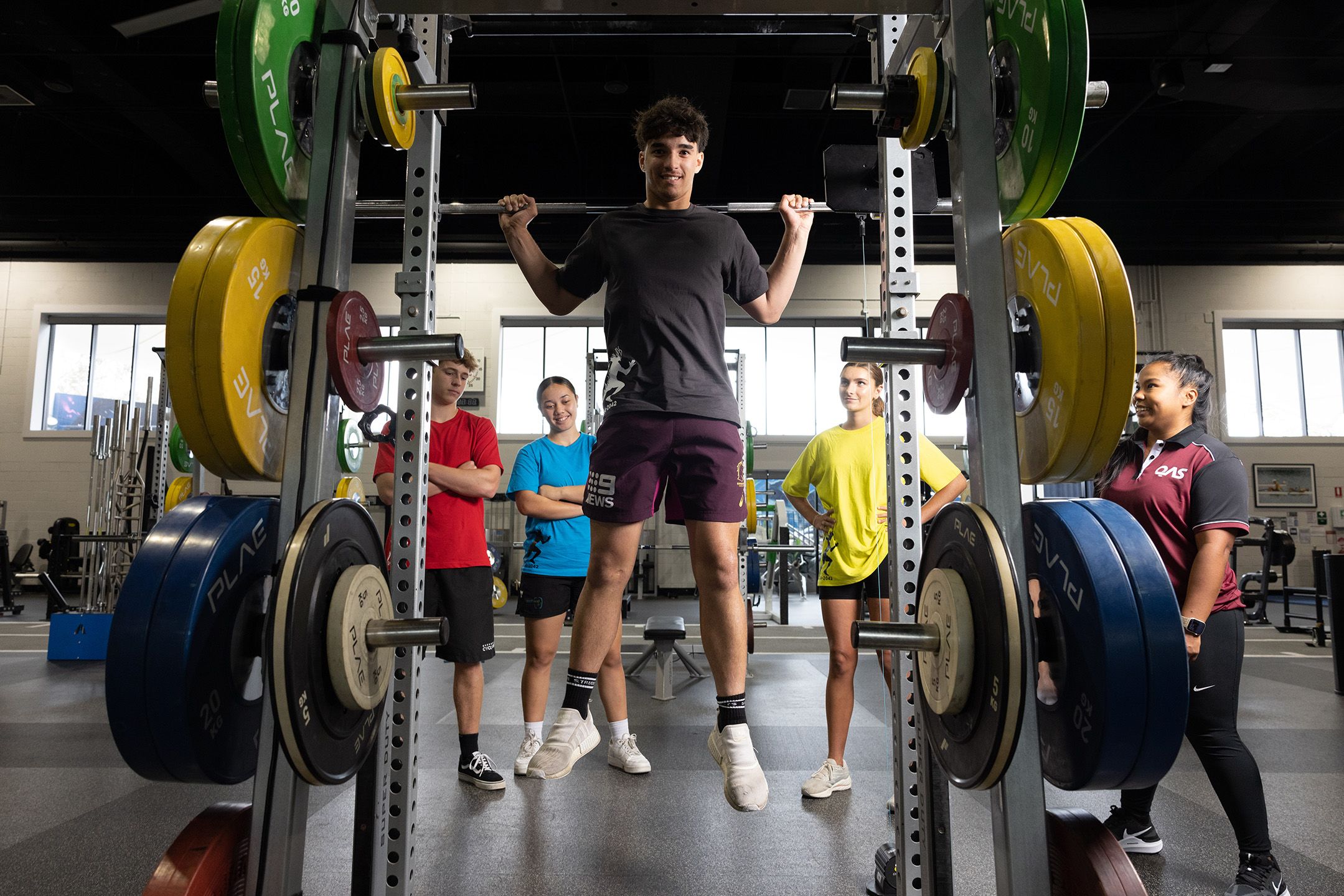 A teenage boy doing a chin up from a barbell, watched on by three fellow students in different coloured tshirts