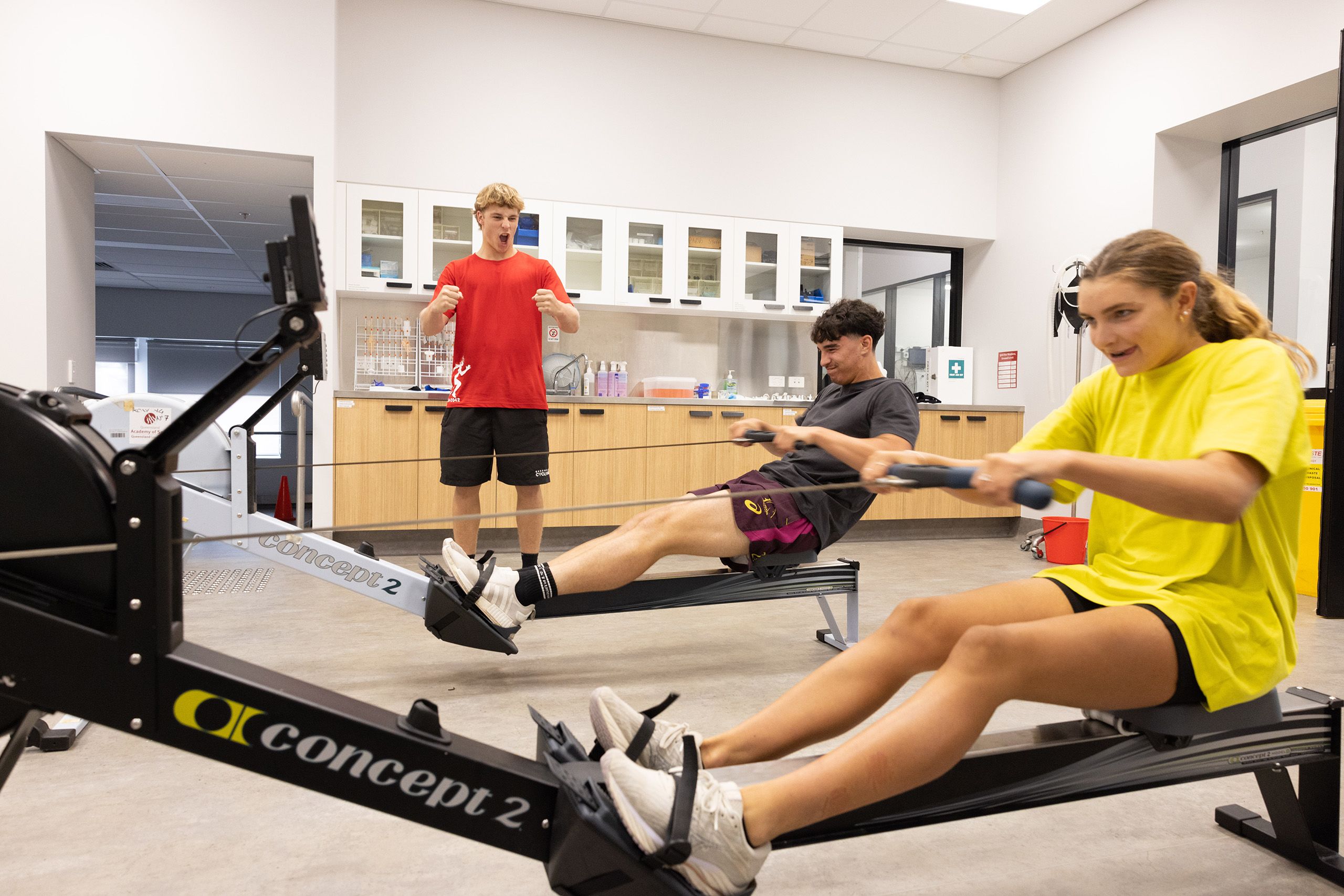Two students, one in a grey t-shirt and one in a yellow t-shirt, compete against eachother on an ergo. They are being cheered on by another student in a red t-shirt