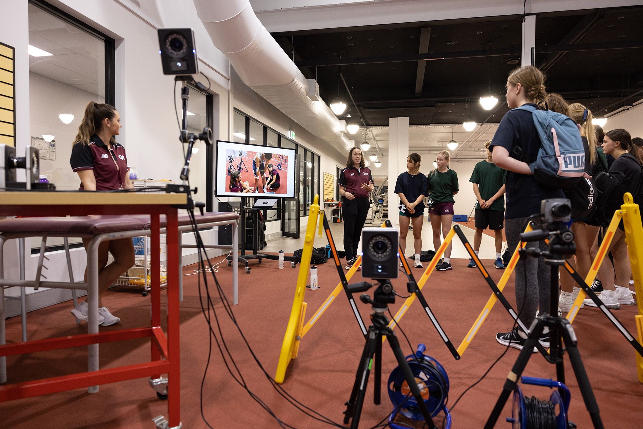A group of high school students gather around a complicated piece of equipment, being addressed by a woman in a maroon polo shirt 