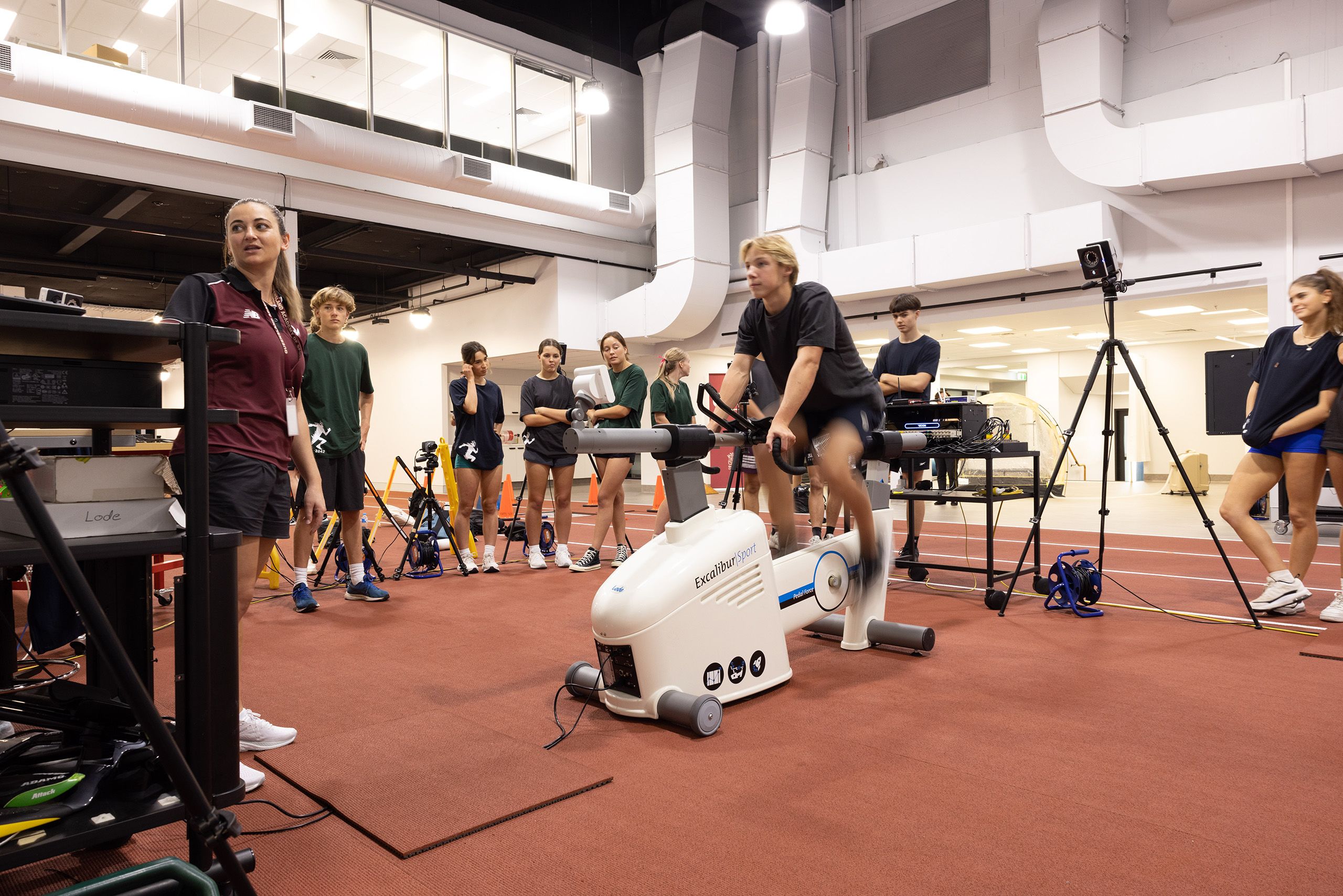 A group of high school students in different coloured t-shirts watches on as a fellow classmate sits on a cycling machine 